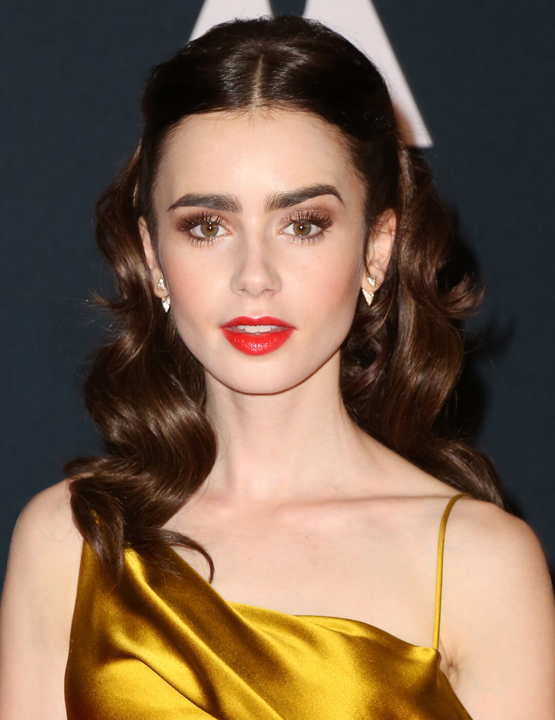 Lily Collins looking elegant in a neutral makeup look and red lips, golden satin dress, and wavy hair