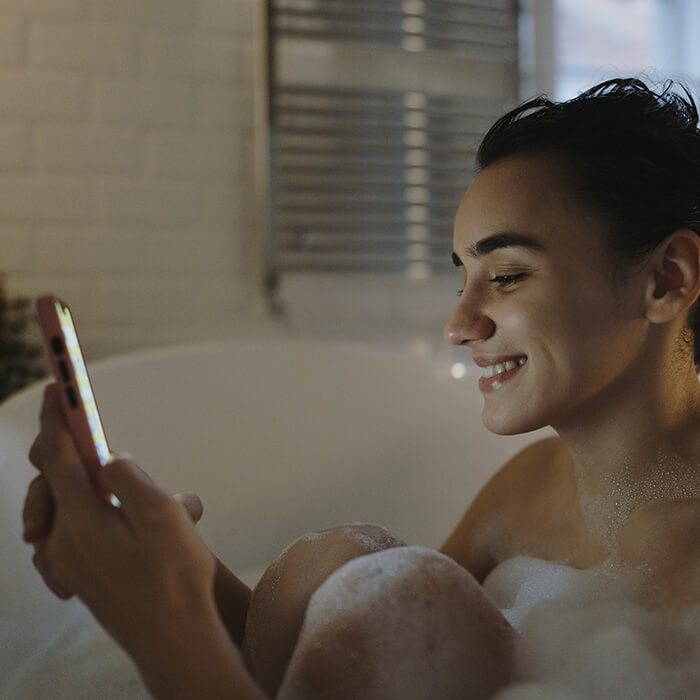 An image of a smiling woman, her hair in a bun, holding her phone above her knees while in the bathtub