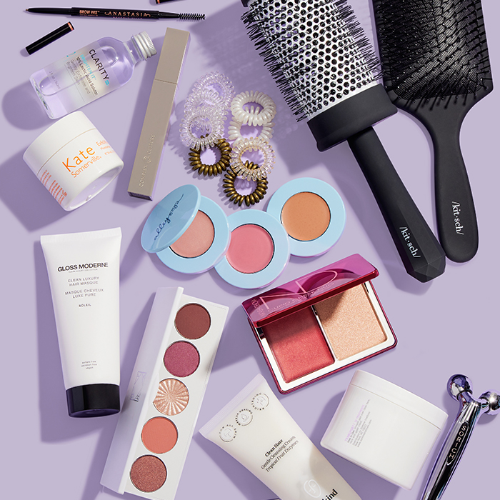 Makeup, skincare, and beauty products and tools from various brands scattered on light purple background