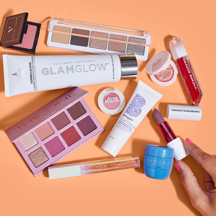 Beauty products from various brands and woman's fingers touching a lipstick against orange background