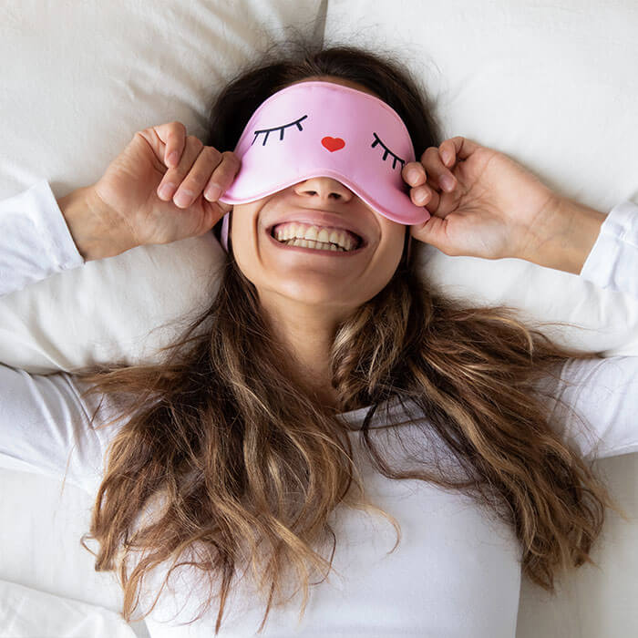 An image of a smiling woman with a cute pink sleeping mask is nestled in bed, wearing a cozy white long-sleeve shirt