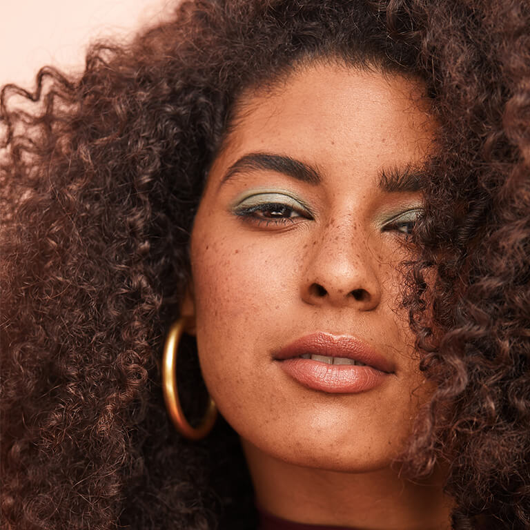 A closeup image of a model with curly hair wearing mint green eyeshadow and gold hoop earrings smiling