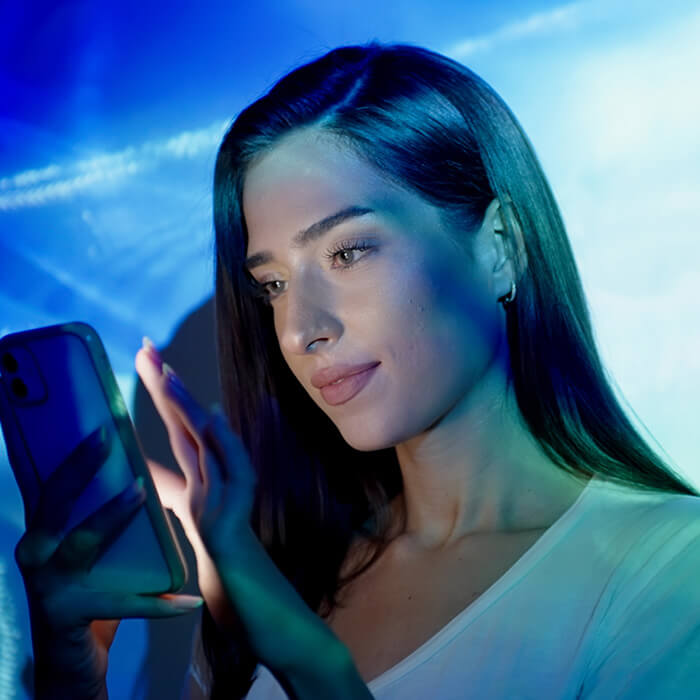 An image of a woman with a subtle smile, glancing at her phone wearing light makeup in a nude lipstick, dressed in a white t-shirt within a room illuminated by various shades of blue