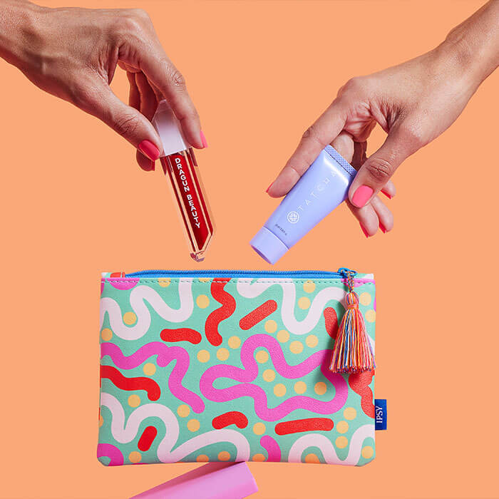 An image with two hands are depicted holding a lipstick and a cream product and endeavoring to place them inside a colorful small bag