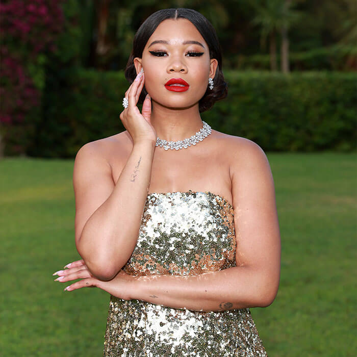 An image of Storm Reid, looking stunning in a gold sequined sleeveless dress, thick black eyeliner, red lipstick, and a set of earrings, rings, and necklaces, with her hair split in the center