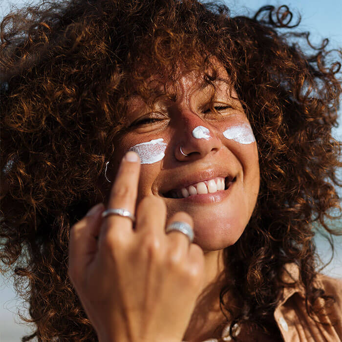 Young woman with curly hair and light brown skin applying sunscreen on her cheeks and nose