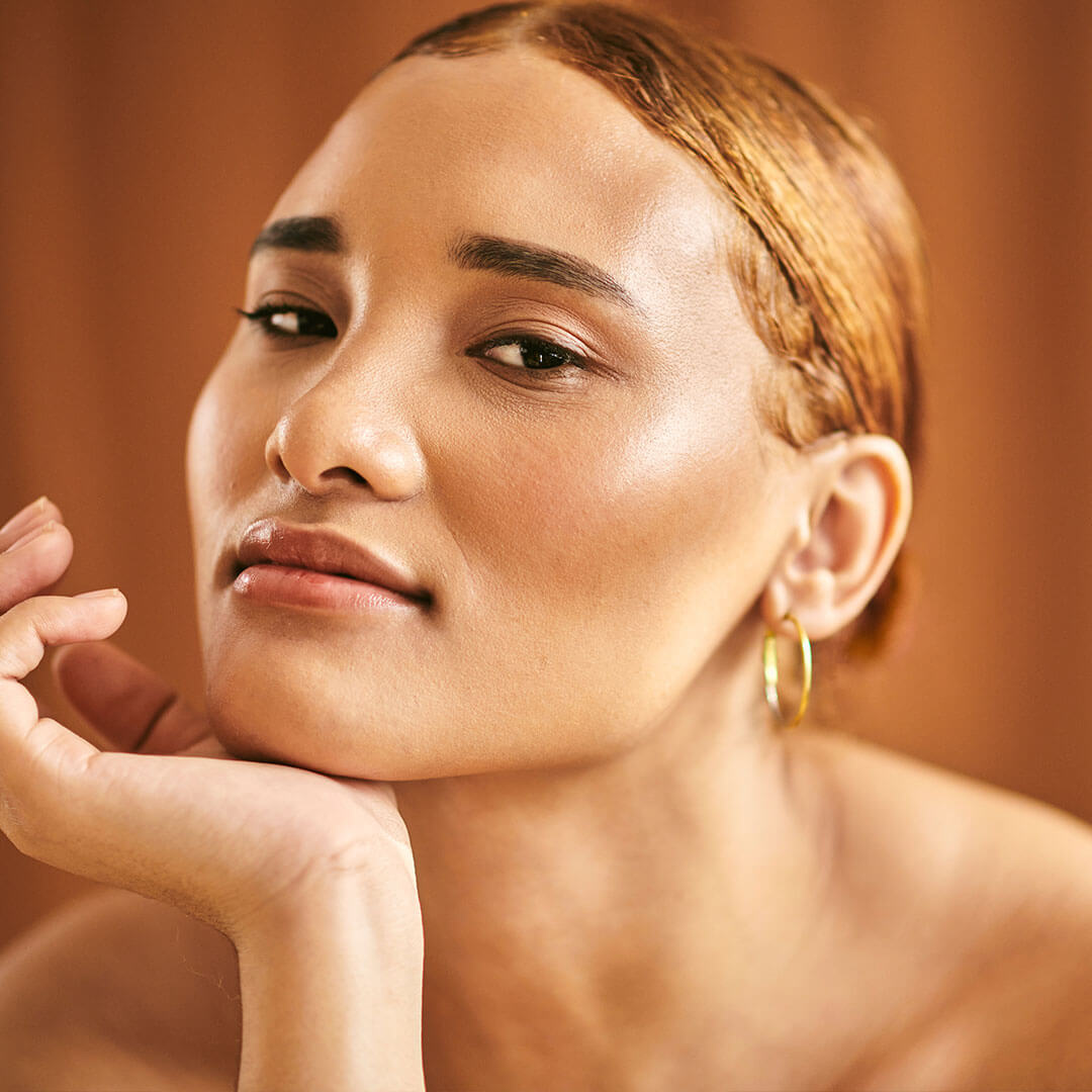 An image of a woman with clear skin, wearing a light touch of makeup with a slick hair parted in half, wearing a gold hoop while her hand is place under her chin against a brown background