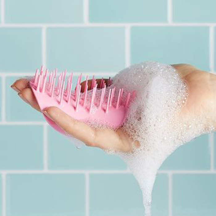 Woman's hand holding a pink scalp massager against teal bathroom tiles
