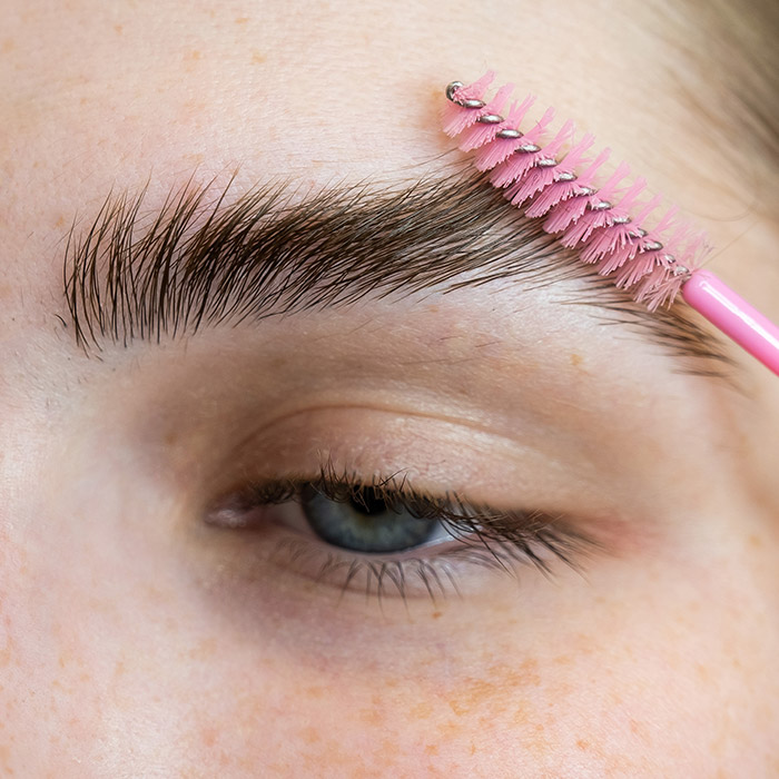 A closeup photo of a model's eye with a pink spoolie brushing her eyebrows