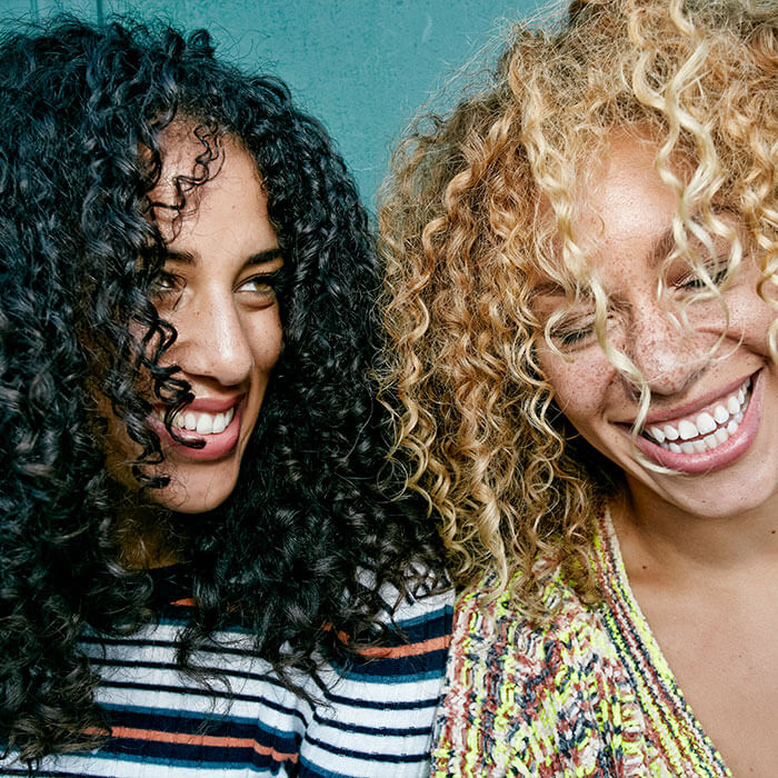 Two women with beautifully textured curly hair, one with dark, tightly coiled curls and the other with blonde, loose curls, showcasing the beauty and vitality of well maintained curly hair