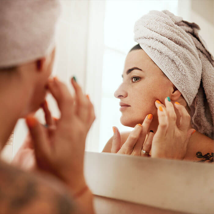 Woman with a towel wrapped around her head, looking in the mirror at her irritated face after applying vitamin C