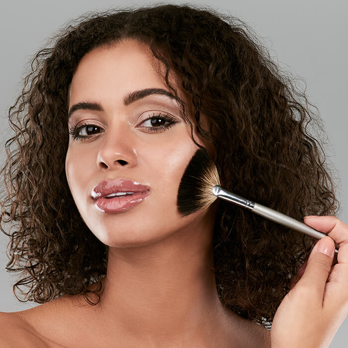 Beautiful woman applying face makeup with a fan brush against grey background