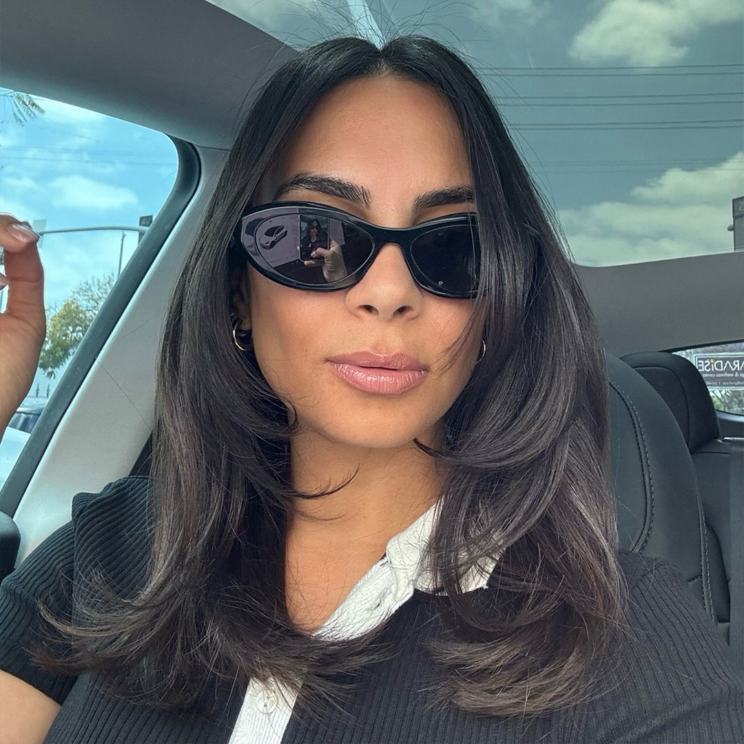 A woman sitting in a car seat, wearing black sunglasses, pouting her lips, and showing off her beautiful black hair