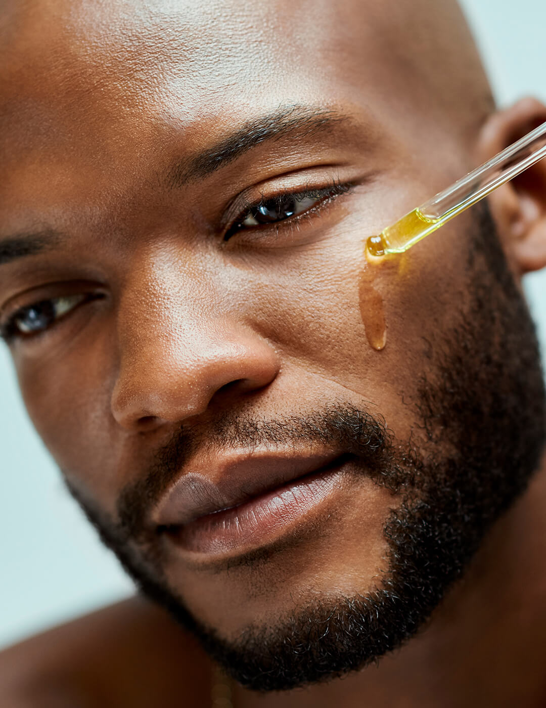 African American applying serum on his face using a dropper. Attractive male model with a beard inside of a room with an isolated studio background. He is wearing a necklace