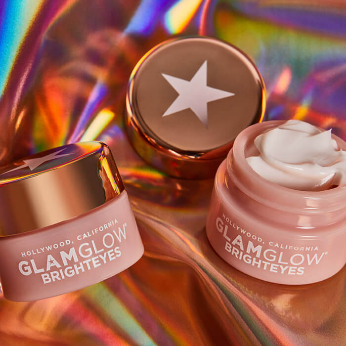 Flat lay image of three jars of GLAMGLOW BRIGHTEYES™ Illuminating Anti-Fatigue Eye Cream against a shimmery cloth background