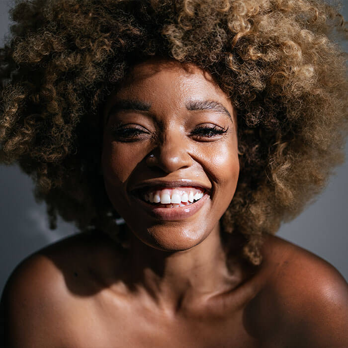 An image of a woman of color with an afro hairstyle is smiling at the camera against a gray backdrop
