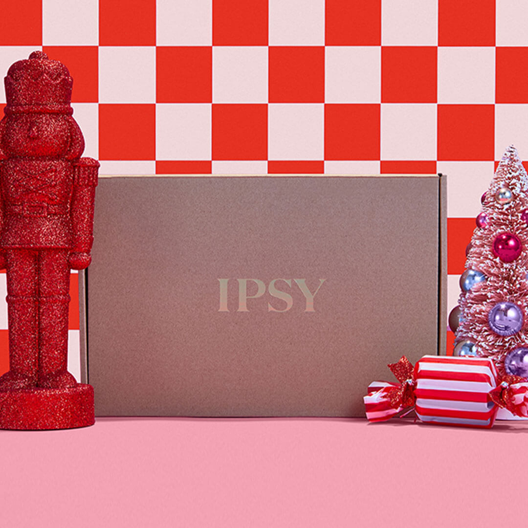 IPSY box against a red and pink checkered background surrounded by holiday-theme props, including a nutcracker and mini christmas tree