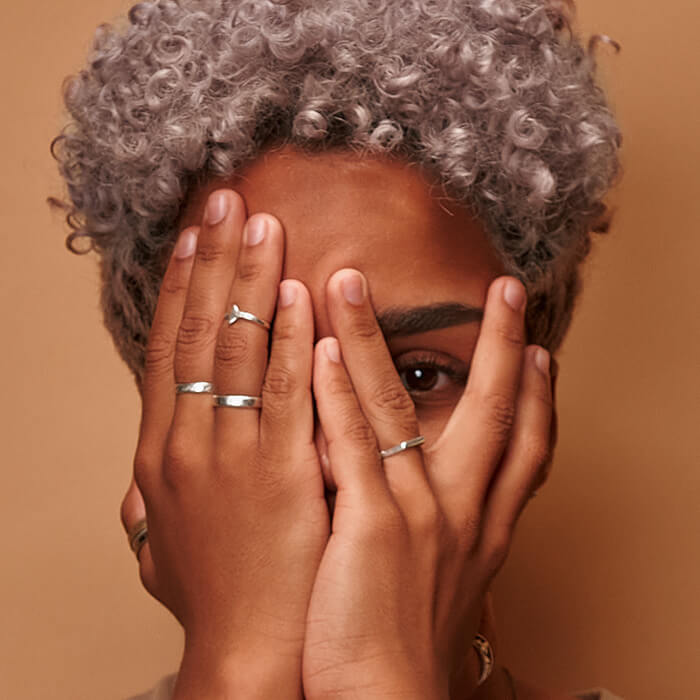 An image of a woman of color with ash blond curly hair partially covering her face with her hands, her fingers adorned with silver rings