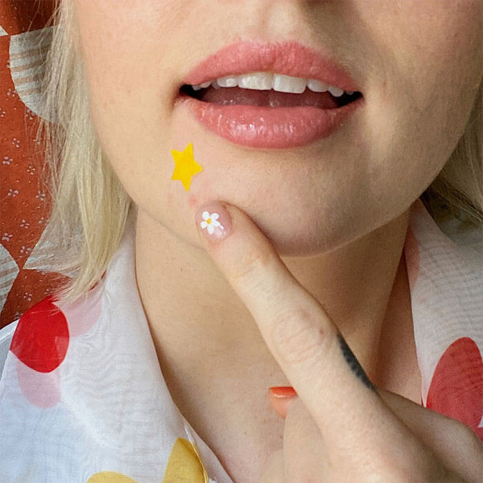 Woman with a star-shaped pimple patch on her face, touching it gently with a finger adorned with daisy nail art