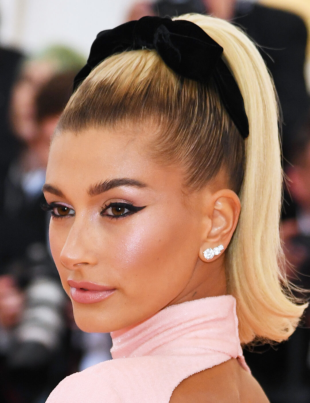 Hailey Bieber rocking a sleek, high ponytail hairstyle, bold winged eyeliner makeup look, diamond earrings, and pink dress