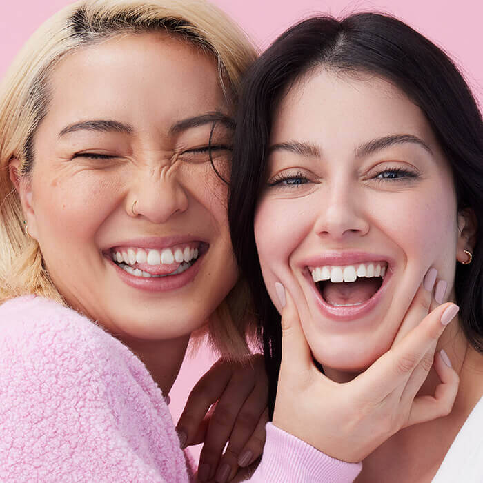 A close-up photo of two women smiling joyfully at the camera while dressed in a white top and a pink sweater, both opting for subtle makeup