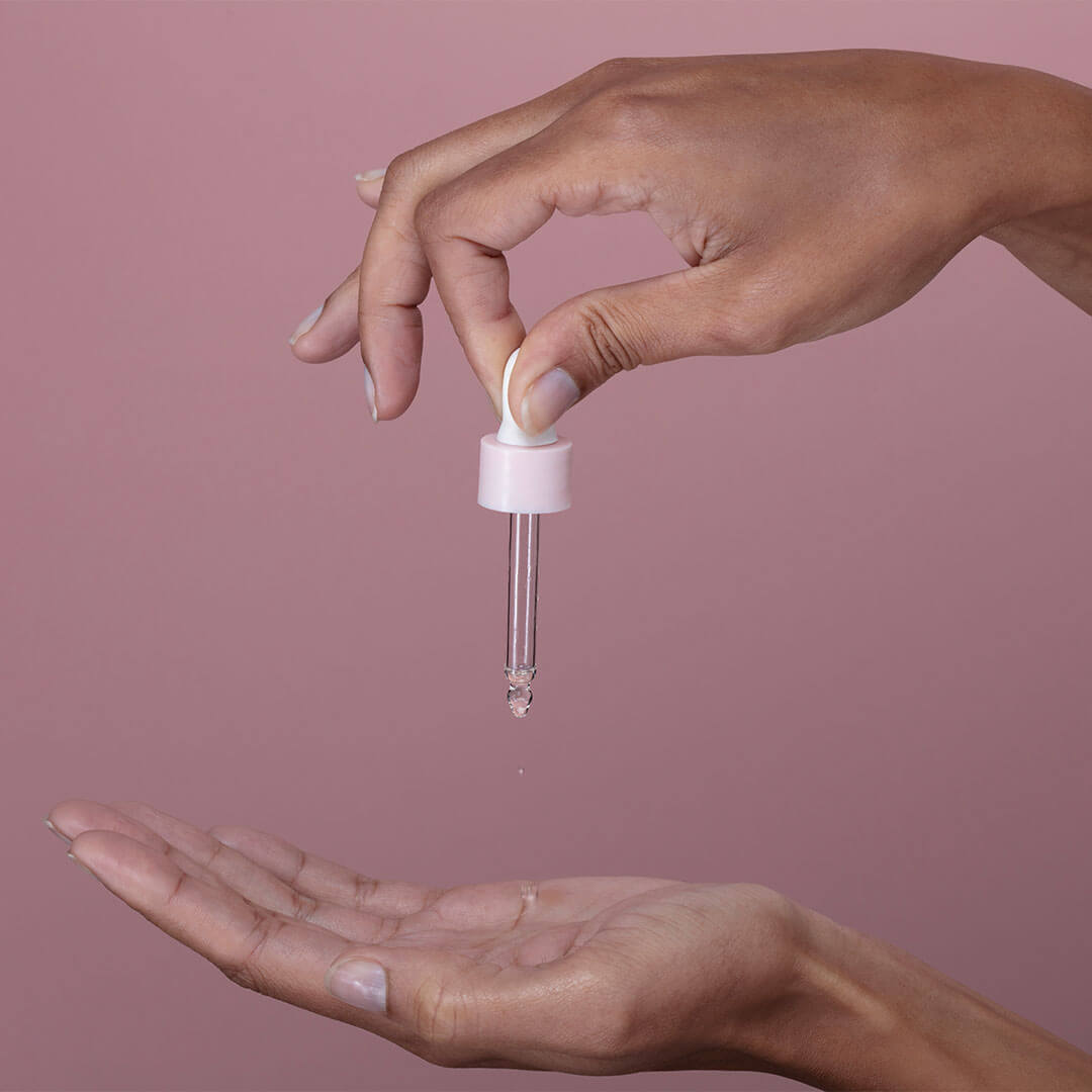 A close-up of hands delicately holding a glass dropper, with a few drops of a clear liquid falling into the palm against a soft pink background