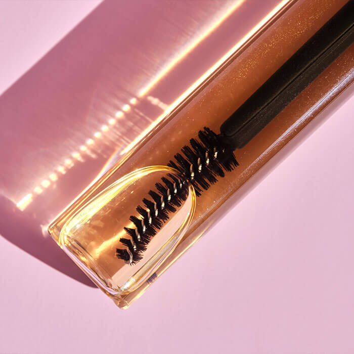 A close-up shot of an eyelash serum applicator brush with fine bristles inside a sleek, transparent tube filled with golden-hued serum