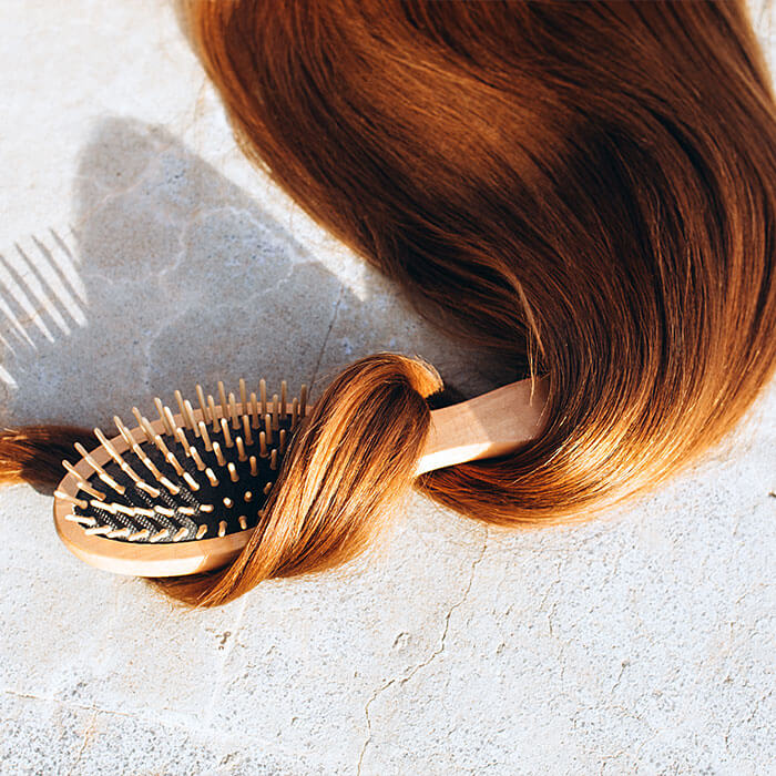 A wooden hairbrush wrapped with long, silky, brown hair on a light concrete surface, emphasizing the importance of maintaining healthy, shiny hair during the summer