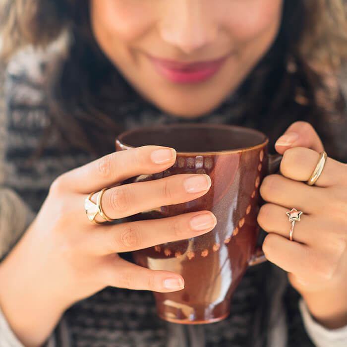 A close-up photo of a woman in a sweater, holding a brown printed mug showing her clean, clear, and polished nails adorned with golden rings in assorted shapes, including stars and triangles