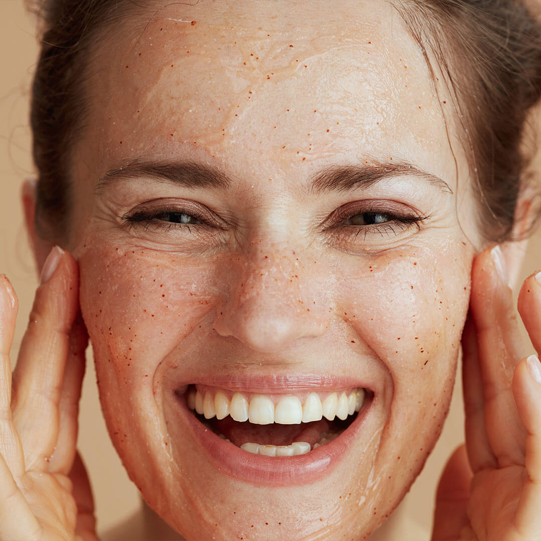 An image of a happy woman, smiling widely infront of the camera while she gently scrubs a face exfoliator onto her skin