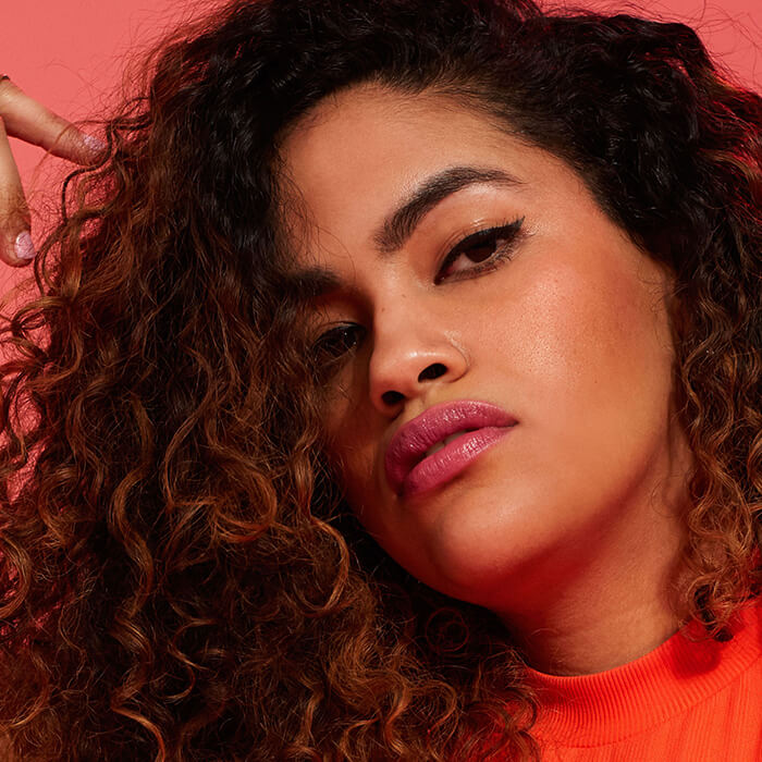 Close-up image of a model with thick, curly hair rocking a natural makeup look wearing an orange top on a red background