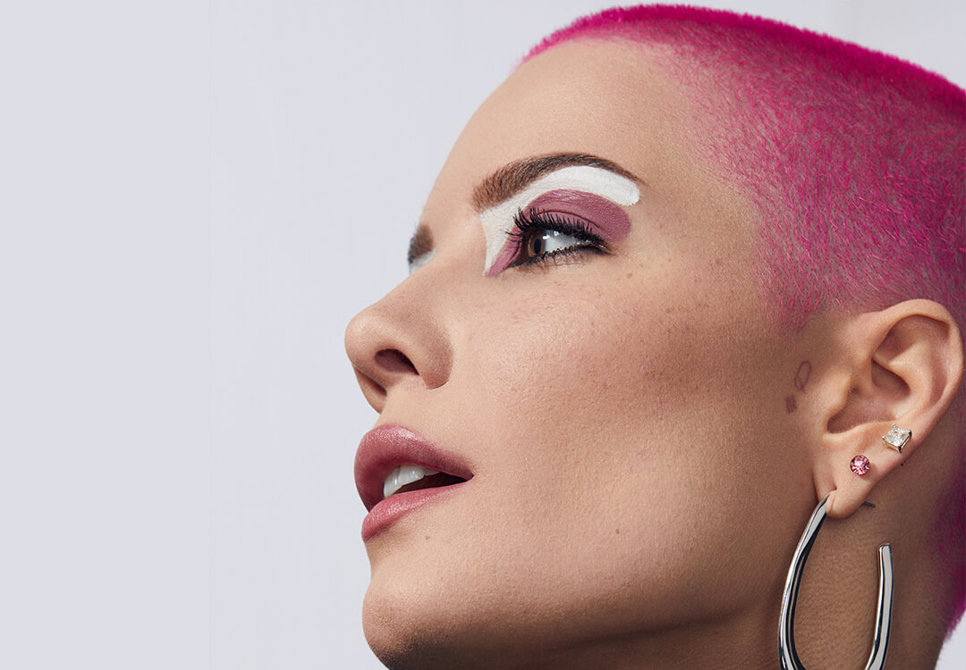 Close-up side profile of Halsey rocking bright pink hair and bold white and pink eyeshadow makeup look paired with a mauve lip and silver dangling earrings