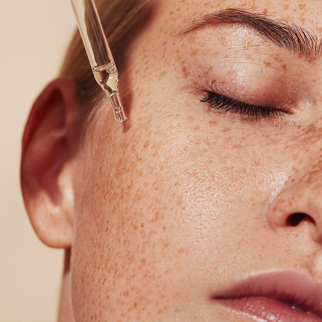 A close-up shot of a woman's face with freckles as she applies liquid serum