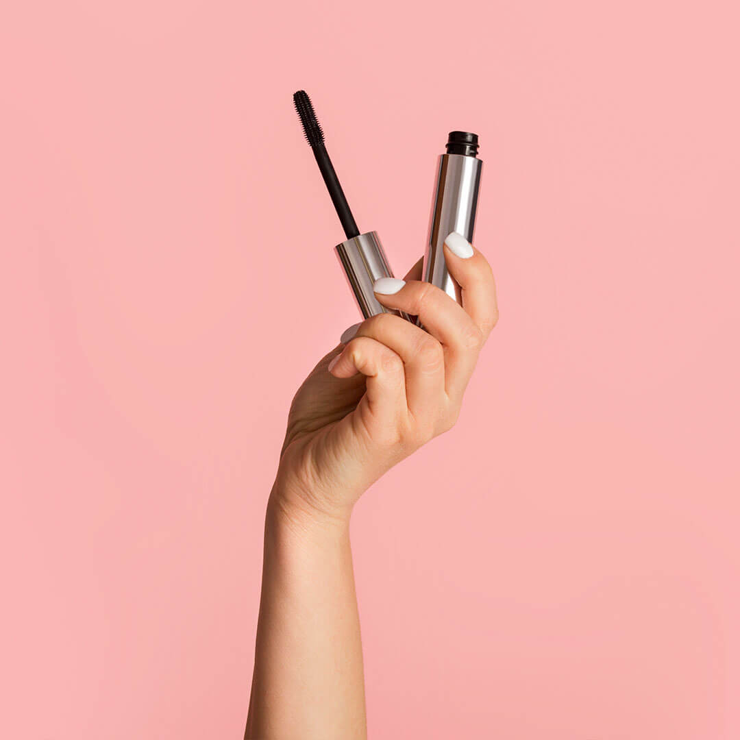 A photo of a hand with white-painted nails holding a mascara tube against a pink background