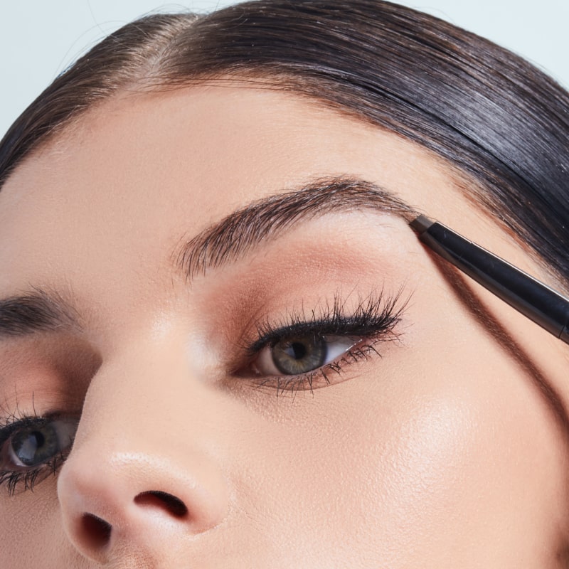 A close-up photo of a woman using a browpencil to fill in her eyebrows while wearing black eyeliner