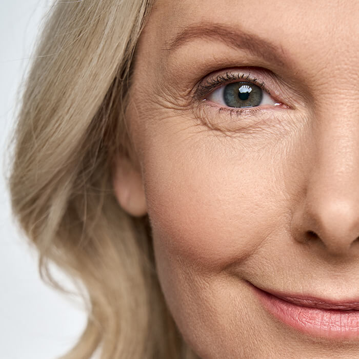 An image of a smiling mature woman in her mid-50s with blonde hair, gazing directly at the camera, revealing a hint of laughter lines around her eye