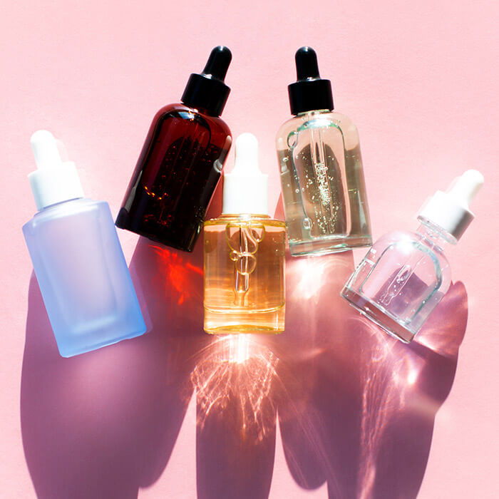 Row of bottles of different shapes, sizes, and colors with beauty products inside against pink background