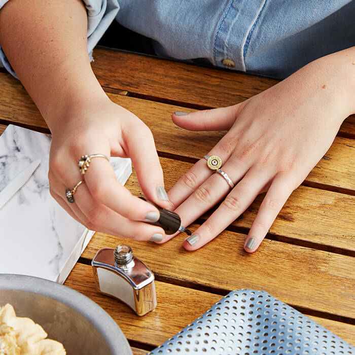 An image of a woman  wearing a denim long-sleeve shirt layered under a stylish leopard-print jacket, wearing several elegant gold rings applying gray nail polish over a wooden table