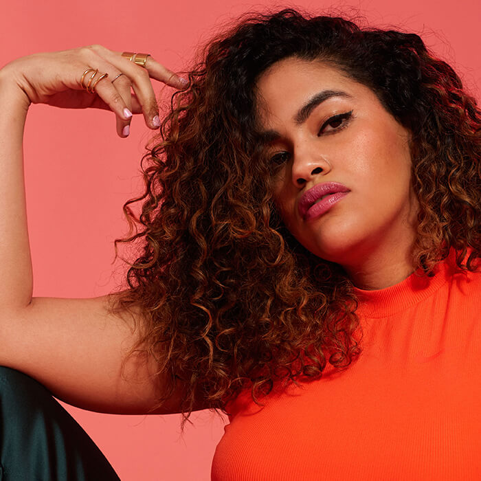 A photo of a woman with curly hair wearing an orange top on a pink background