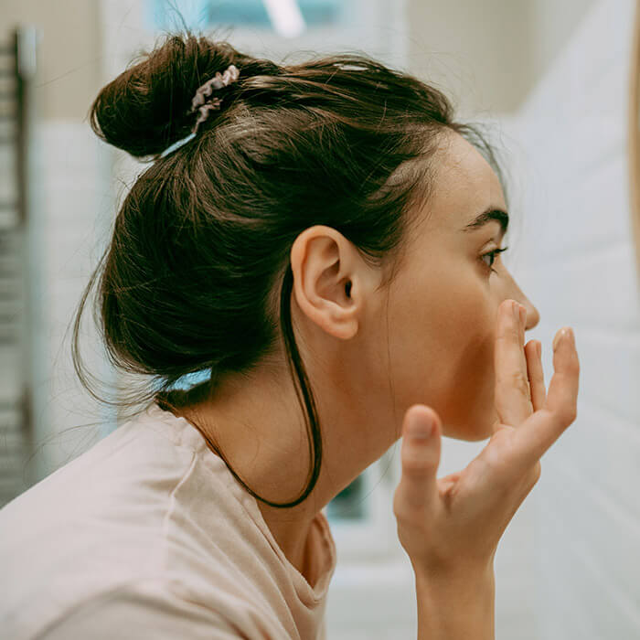 A photo of a woman applying moisturizer on her face in front of a mirror in the bathroom