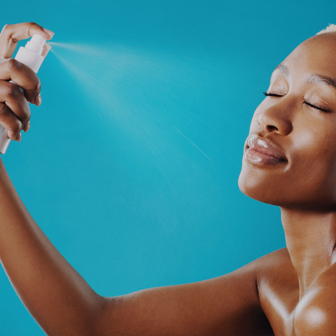 An image of a young woman of color, misting a setting spray onto her face