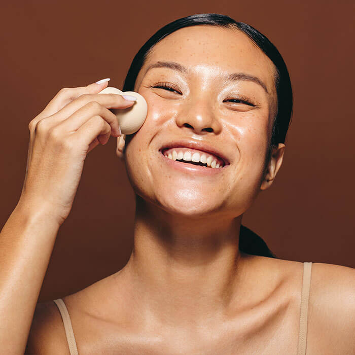 A picture of a happy woman applying a mixture of foundation and moisturizer with a beauty sponge to her face