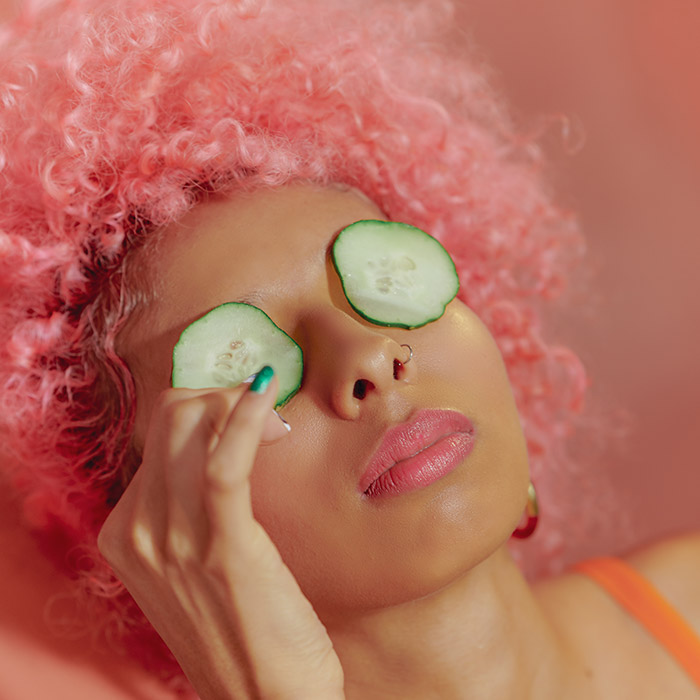 Close-up image of a model wearing an orange tank top  and pink curly wig with cucumbers on top of both eyes 