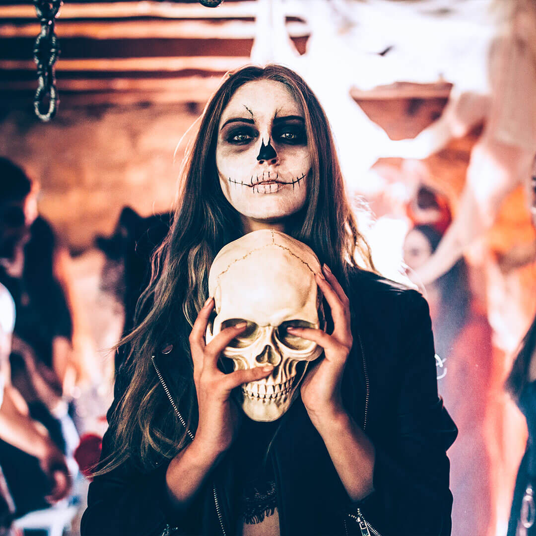 A woman in striking skull face paint holds a realistic human skull, standing amidst a dimly lit Halloween party, surrounded by other costumed figures in the background