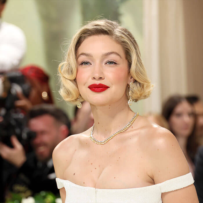 A photo of Gigi Hadid showing off her short blond hair, bright red lipstick, white off-the-shoulder dress, and diamond gold necklace