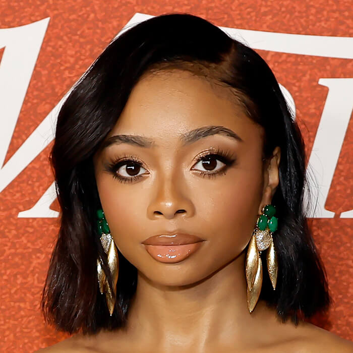 An image of Skai Jackson donning medium-length black hair while sporting a natural-toned lip color, alongside striking jade and golden drop earrings