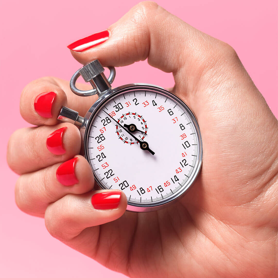 A hand with bold red nail polish holds a silver stopwatch, set against a bright pink background