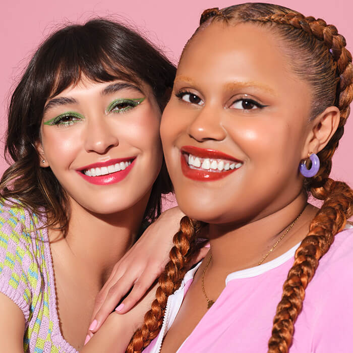 A close-up image of two women smiling towards the camera showcasing current makeup trends as they smile at the camera, both elegantly dressed—one in a pink top and the other in a knitted top with vibrant neon prints