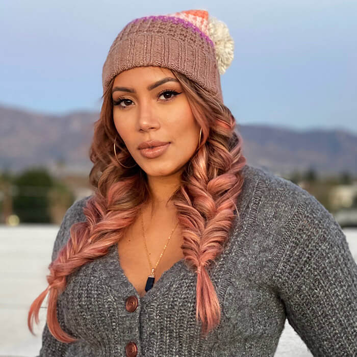 Close-up image of Cynthia Alvarez rocking rose gold hair, dark gray cardigan, and rose beanie and posing against mountain and tress background