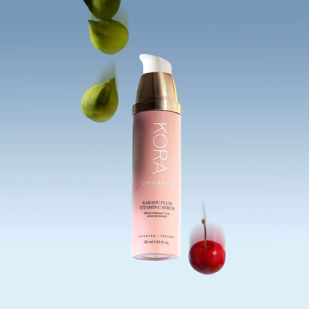 A pink and gold bottle of KORA ORGANICS Kakadu Plum Vitamin C Serum stands against a serene blue backdrop, with a fresh green leaf and a bright red fruit